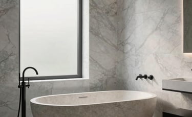 A high-end, luxury bathroom in a Northern European / Dutch home. The scene features large marble wall tiles in light gray and white, a freestanding stone bathtub, and minimalist matte black fixtures. Soft, diffused morning light enters through a frosted window. The composition is clean and architectural, highlighting the exquisite craftsmanship and serene atmosphere.