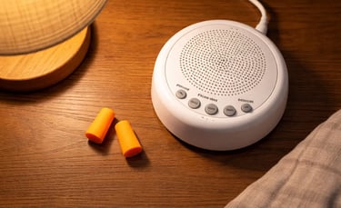 White noise machine and orange foam earplugs on a wooden bedside table with a warm lamp for better sleep.