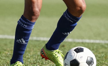 Close-up of a young soccer player's feet, diverse origin, performing a technical dribble with a modern soccer ball. European training facility background. The Total Football Academy logo is subtly placed on the player's sock. Bright daylight, dynamic motion blur.