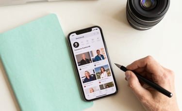 A top-down professional flat lay of a content planning session in a bright office in a Spanish-speaking region. The scene includes a mint green notebook, a modern smartphone showing social media feeds, and a hand holding a pen. The aesthetic is clean and organized with beige and matte white surfaces.