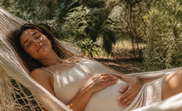 A serene pregnant woman with dark hair rests peacefully in a hammock outdoors, gently cradling her baby bump in the warm