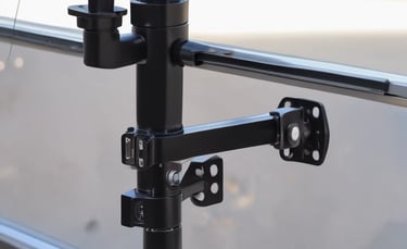 A small, modern teardrop trailer with a white body and bright yellow accents is parked on a grassy area. The trailer features a curved shape, a small window with a yellow frame, and a circular company logo displayed on its side. The trailer is attached to a vehicle that is partially visible.