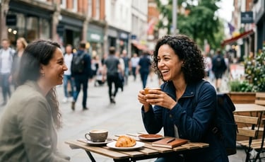Duas mulheres sorrindo e conversando em um café na calçada, com café e croissants, em uma rua.