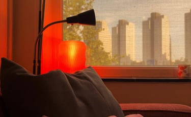 Cozy living room corner with a red glowing lamp, plush pillows, and a window view of city skyscrapers at sunset.