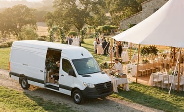 A clean white delivery van parked next to an elegant outdoor wedding tent setup, warm golden hour sunlight, soft editorial style