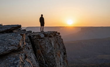 A wide-angle landscape shot of a person standing on a rugged North American cliff at dawn, looking towards a vibrant gold sun rising over the horizon, clean and modern photography style, soft morning light.
