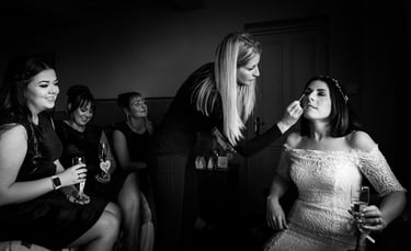 Professional makeup artist applying eye makeup to a bride in a lace wedding dress with bridesmaids.