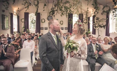 Smiling bride in floral crown and groom at an indoor rustic wedding ceremony with greenery decor.