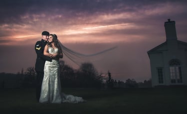 Military wedding couple embracing at sunset with a flowing bridal veil and manor house background.