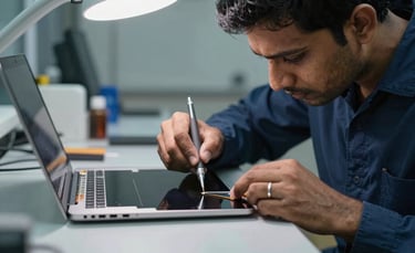 A close-up photograph of a South Asian technician in a clean, modern workshop in Thane, carefully replacing a thin laptop screen using precision tools under bright, focused lighting. The atmosphere is professional and high-tech, featuring a palette of dark blue and light grey.
