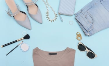 Flat lay of women's fashion accessories including heels, jeans, jewelry, and makeup on a blue background.