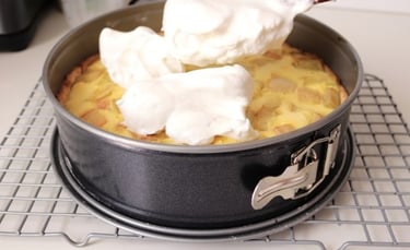 Spreading fluffy white meringue over a freshly baked apple tart in a springform pan on a cooling rack.