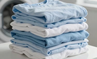 A close-up photograph of a neat stack of freshly folded cotton clothes in soft shades of light blue and white. The setting is a bright, modern North American laundry room with clean surfaces and natural light.