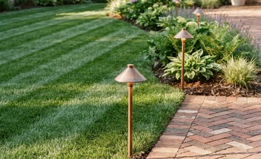 Crisp outdoor photography of a perfectly striped green lawn meeting a brick patio, with neat copper-accented landscape lighting fixtures casting warm morning light along the edge, 35mm.