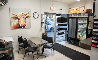 Indoor seating area at North Port Subs featuring a colorful cow painting, dining table, and beverage cooler.