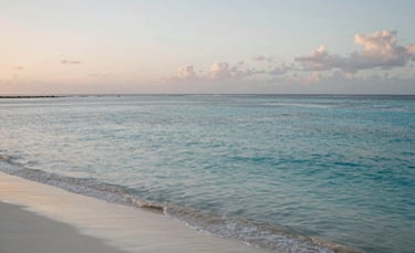 A peaceful coastline in the Caribbean / Anglophone Caribbean at dawn, with calm turquoise waters and soft white sand. The lighting is gentle and serene, emphasizing a sense of tranquility and restoration.