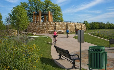 Cyclists ride on a paved trail near a stone pavilion on the San Antonio River Trail.