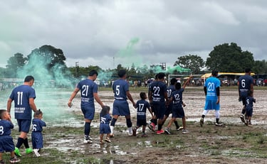 Soccer team and children walk across a muddy field with green smoke flares under a cloudy sky.