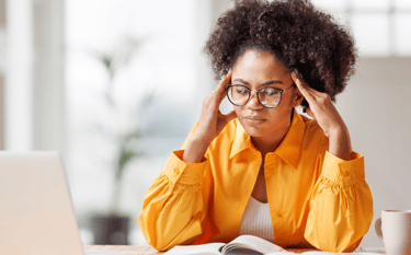 a woman in glasses and yellow shirt is sitting at a table with laptop and notepads looking stressed