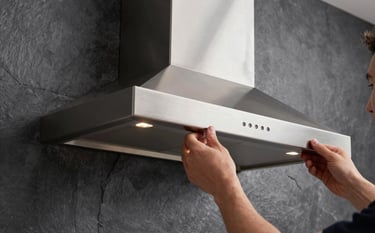 A close-up of a sleek designer kitchen chimney being mounted on a dark slate grey wall. Professional hands are visible ensuring precise alignment. Premium lighting highlighting the brushed metal finish.