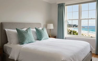 A luxurious holiday home bedroom on the Victorian coast. The bed is perfectly made with crisp white linen and light teal decorative pillows. Through the window, the Australian coastline is visible. The room feels inviting and impeccably clean. Bright, airy, and sophisticated photography.