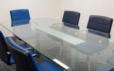 A high-angle shot of a clean, modern conference room in a European / French business district. A glass table reflects soft lighting. The room features royal blue and dark navy blue accents. Minimalist and professional photography style.