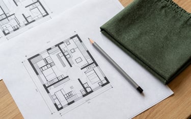 A top-down professional photograph of architectural blueprints spread out on a clean wooden desk. A silver drafting pencil and a swatch of deep olive green fabric sit nearby. Soft, natural morning light creates a clean, modern aesthetic.