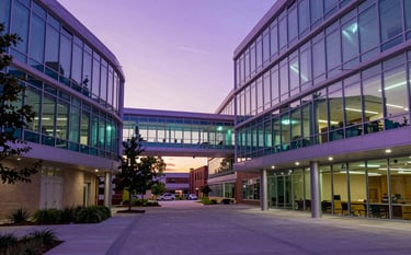 Photography of a North American / US campus courtyard at sunset. Architectural lighting in purple and electric cyan illuminates the glass facades of modern buildings.