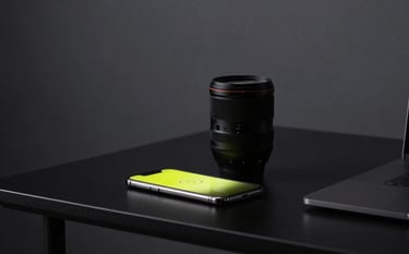 A minimalist, modern workspace with a matte black desk. On the desk sits a high-end camera lens and a glowing smartphone screen reflecting a neon yellow brand logo. The background is a soft-focus dark charcoal studio wall.