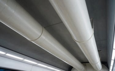 Architectural photography of the Riyadh Metro interior, focusing on the massive exposed ductwork systems installed along the ceiling. The composition is a dynamic low-angle shot, with cool off-white surfaces and soft slate blue industrial accents.