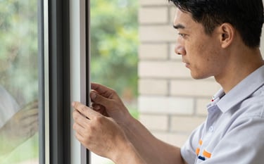 A close-up of a professional in a clean uniform inspecting property window frames. The background shows a soft off-white brick wall and soft green foliage. The focus is sharp, highlighting a meticulous and trustworthy service.