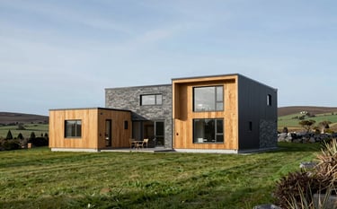 A spacious, two-story contemporary modular family residence in a lush Irish green landscape. The house features mixed textures of wood and slate gray metal, with a professional architectural photography style under a clear light blue sky.