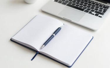 A professional and clean top-down view of an organized desk with a notebook, a pen, and a laptop. The lighting is soft and natural. The color palette features whites and soft blues (#A0C2D9), reflecting a clear and concise work environment.