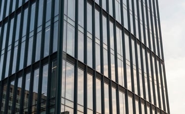 A professional architectural shot of a sleek glass office tower in a metropolitan area. The facade reflects a mist white sky. The structure uses dark charcoal green steel beams for a modern, industrial-minimalist look. Shot at sunset to emphasize transparency.
