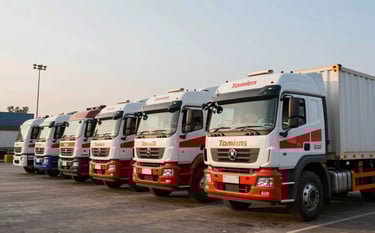 A fleet of modern heavy-duty cargo trucks with clean white and red branding lined up at a logistics terminal in India during the golden hour. The setting is a professional South Asian transport hub with clear skies and efficient industrial vibes.