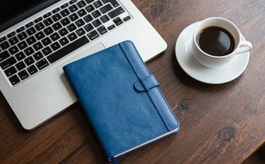 A top-down view of a professional's desk with a laptop, a steel blue notebook, and a clean cup of coffee. The setting is clean and organized, conveying expertise and trustworthy management of financial data.