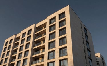 A low-angle, wide shot of a modern architectural residential building with clean lines and large glass windows. The building is made of soft off-white stone and is illuminated by the warm, golden glow of a setting sun. The sky is a deep, sophisticated dark charcoal.