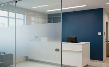 Clean and professional photography of a modern medical clinic reception in Brazil. The scene shows glass partitions with high-quality frosted vinyl branding, soft natural lighting, and a minimalist design in shades of dark blue and white.