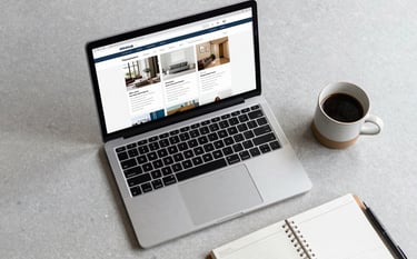 A top-down view of a designer's workspace featuring a modern laptop showing a clean website dashboard, a ceramic cup of coffee, and a minimal planner on a pale pebble gray desk.