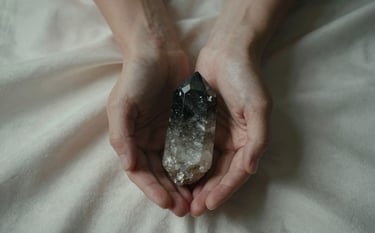 A top-down photographic view of two hands gently holding a charcoal gray quartz crystal over a soft off-white silk surface. The lighting is low and moody, casting soft shadows, evoking a sense of ancient spiritual wisdom.