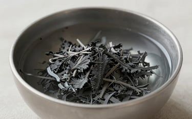 A close-up photograph of a silver gray bowl filled with pure water and charcoal gray herbs. The background is soft off-white linen, symbolizing purity and healing through traditional spiritual means.