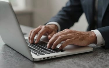 A close-up photograph of a professional's hands working on a silver laptop in a refined North American / European interior. The environment features muted light grey textiles and charcoal slate accents. The lighting is soft, overhead, and professional.