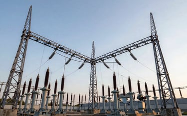 A wide-angle professional photograph of a massive industrial electrical substation. Steel lattice towers and large porcelain insulators stand tall against a Soft Mist Blue sky. The composition is symmetrical, highlighting engineering precision. Lighting is crisp morning sun, emphasizing the Deep Navy Blue metallic structures.