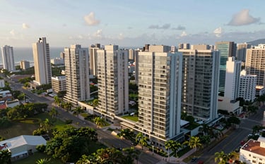 A cinematic aerial photograph of a new real estate development project in a South American / Brazilian coastal city. Modern apartment buildings with clean glass facades are integrated with lush green landscaping. Morning light. Colors: slate blue, soft white.