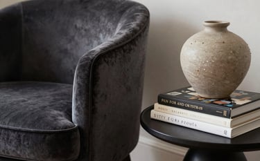 A close-up photograph of a beautifully curated interior in a North American home. A refined charcoal velvet chair sits next to a side table with sophisticated art books and a sand-colored ceramic vase. The mood is premium and artistic with soft, natural depth.
