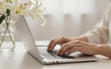 Close-up of hands elegantly typing on a modern, slim keyboard next to a delicate glass vase of white lilies. The lighting is soft and airy, emphasizing a refined feminine touch and a high-end personal brand aesthetic using ivory, cream, and soft bronze tones.