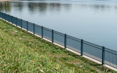 A clean, well-maintained shoreline with a modern security fence. The grass is neatly trimmed, and the water's edge is free of debris. Professional craftsmanship is evident in the fencing. Brand colors #4F7F8F and #A0CBD9 reflected in the lake and metalwork.