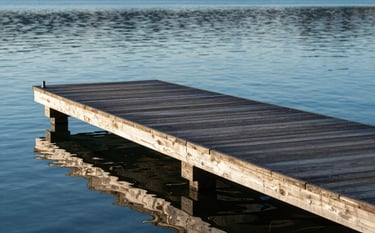 A high-end custom wooden dock with precision joinery extending into a calm blue lake. The timber has a professional, dark protective finish. Morning sun creates soft reflections on the water. The scene incorporates brand colors #1B3A4B and #4F7F8F in the shadows and wood tones.