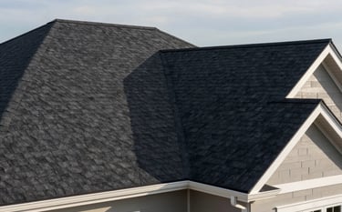 A high-angle, professional photograph of a freshly installed charcoal asphalt shingle roof on a modern North American home. The sunlight highlights the precision and clean lines of the craftsmanship, with a soft blue-grey sky in the background and white architectural accents.