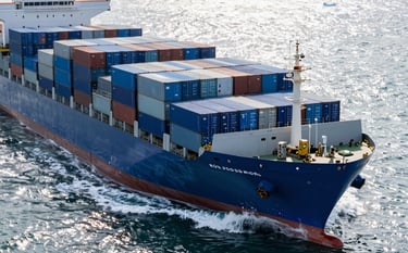 Professional high-angle shot of a massive cargo vessel at sea, stacked with steel blue and deep navy shipping containers, bright daylight reflecting off calm ice white waves, modern and efficient atmosphere.